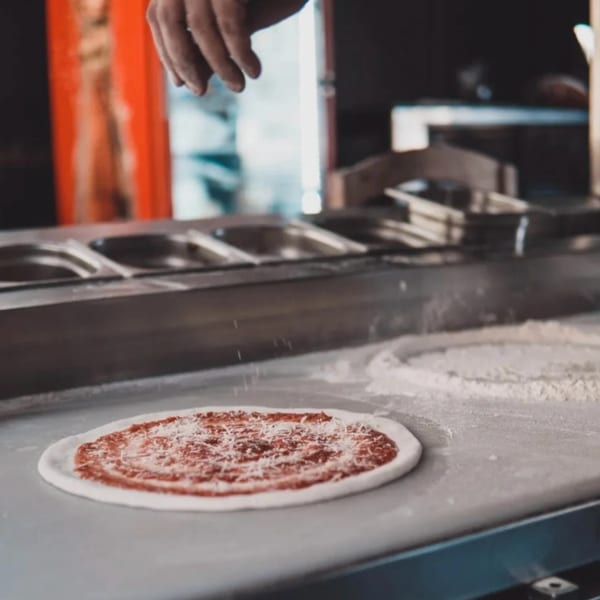 A hand sprinkles cheese onto a Ruszoni's pizza with red sauce on a white dough base.