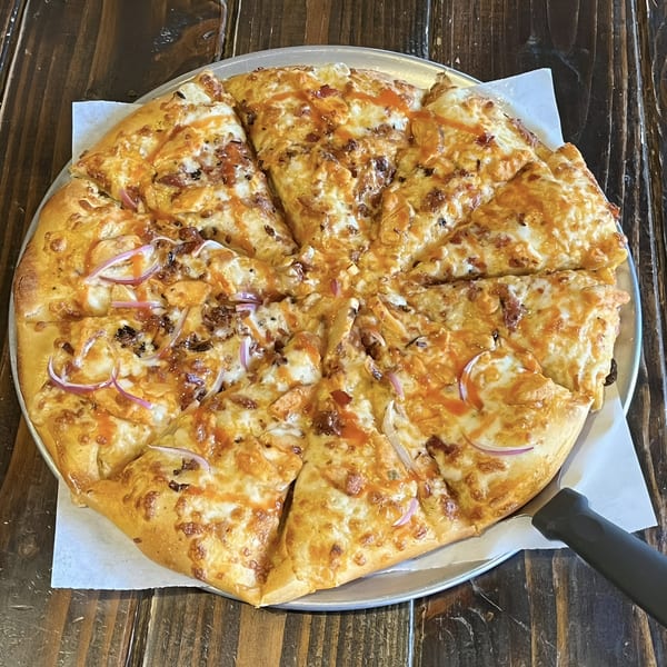 A Ruszoni's Pizza with chicken, red onions, and a drizzle of sauce, on a metal pan.